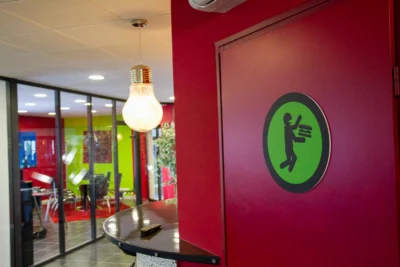 Un espace de bureau moderne aux murs rouges et verts est mis en valeur, avec une porte arborant un pictogramme humoristique représentant une personne mangeant un énorme burger, une suspension en forme d’ampoule lumineuse, et une salle de réunion vitrée visible en arrière-plan. - Agrandir l'image 19 sur 28, fenêtre modale