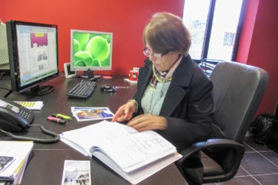 Une femme, vêtue d’une veste sombre et portant des lunettes, est assise à son bureau dans un bureau aux murs rouges, concentrée sur un grand cahier ouvert, entourée de documents, surligneurs et d’un téléphone, avec deux écrans d’ordinateur affichant des pages de travail en arrière-plan. - Agrandir l'image 20 sur 28, fenêtre modale